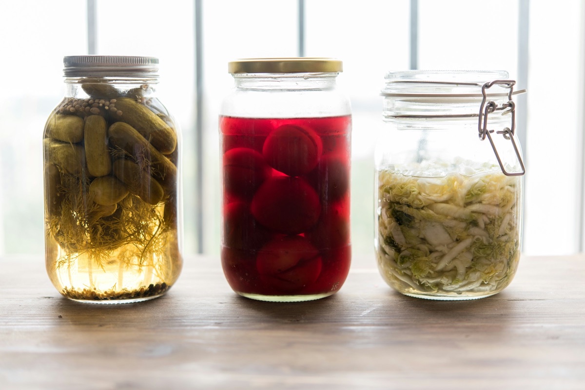 Drei Gläser mit eingelegtem Gemüse auf einem Holztisch, darunter Gurken und Sauerkraut