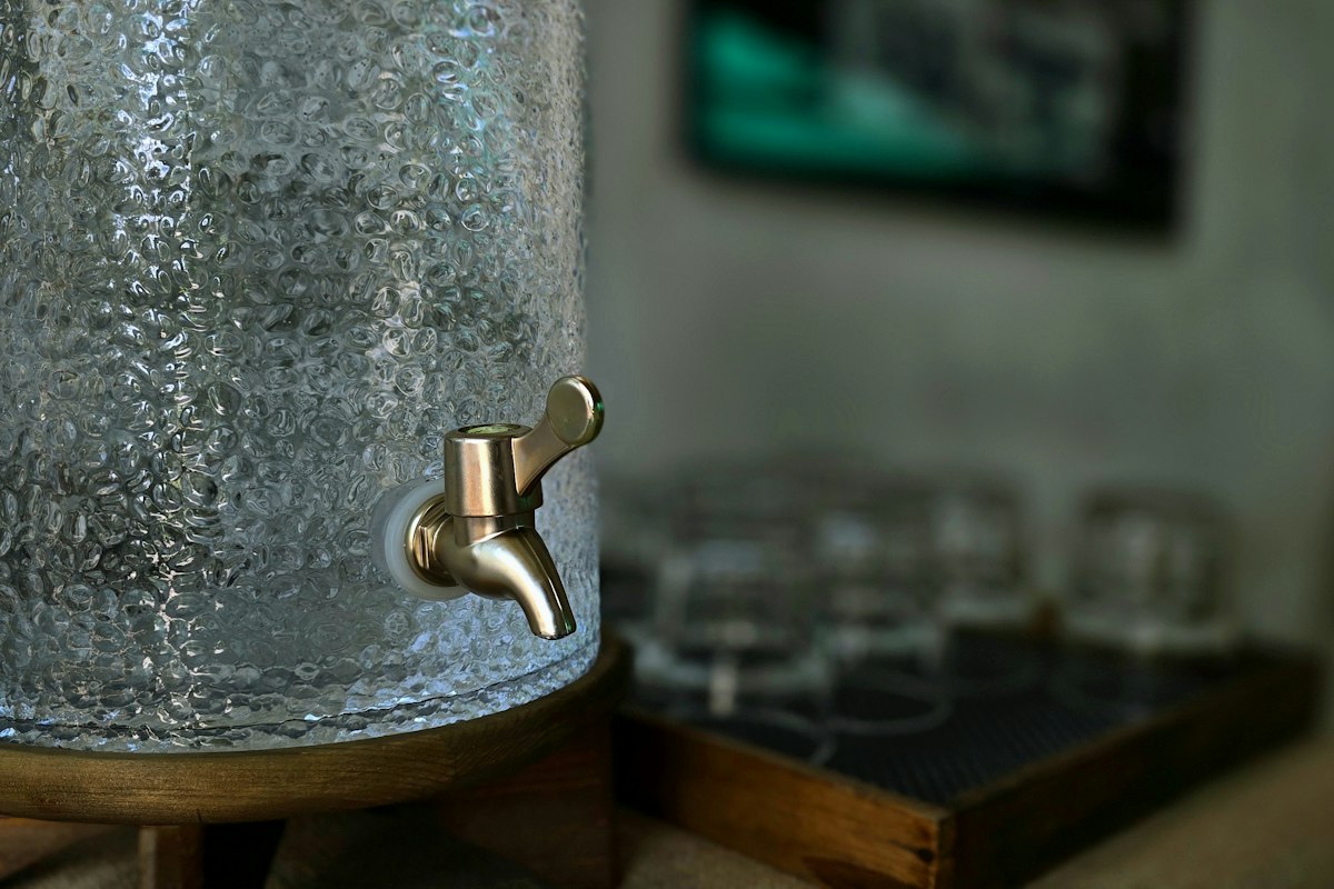 Glasbehälter mit Zapfhahn, im Hintergrund Gläser auf einem Tablett