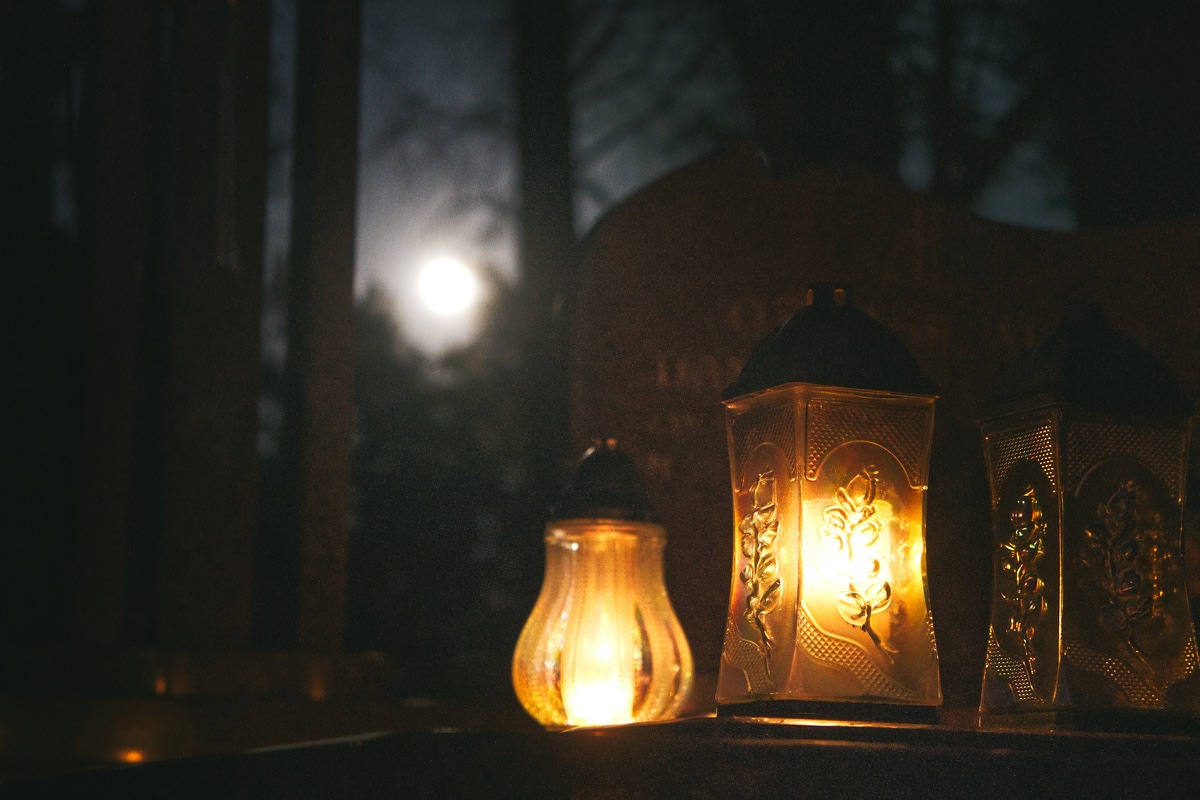 Grablaternen leuchten nachts auf einem Friedhof, dahinter steht der Vollmond zwischen kahlen Bäumen