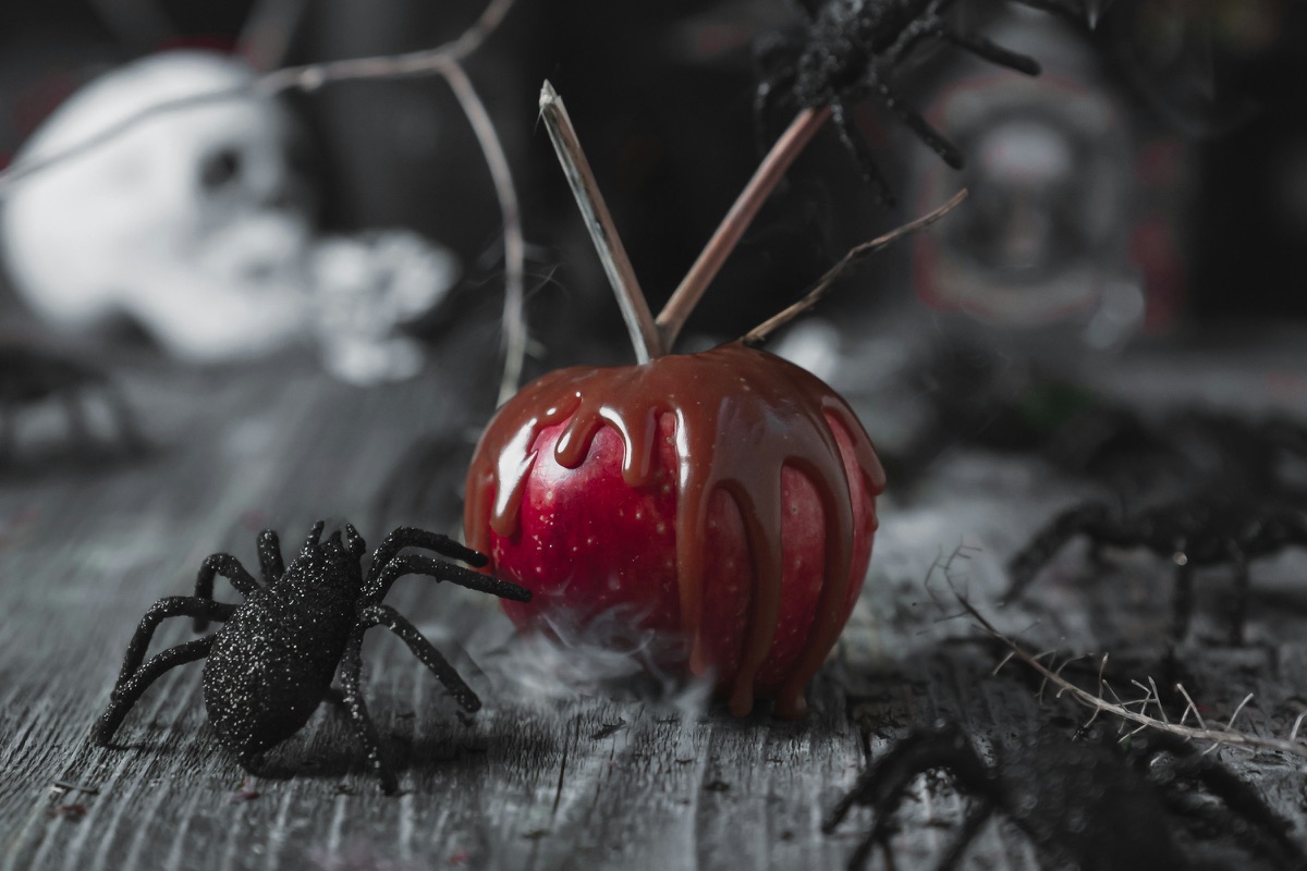 Ein kandierter roter Apfel auf einem alten Holztisch, daneben schwarze Spinnen, im Hintergrund ein Totenschädel und Rauchschwaden