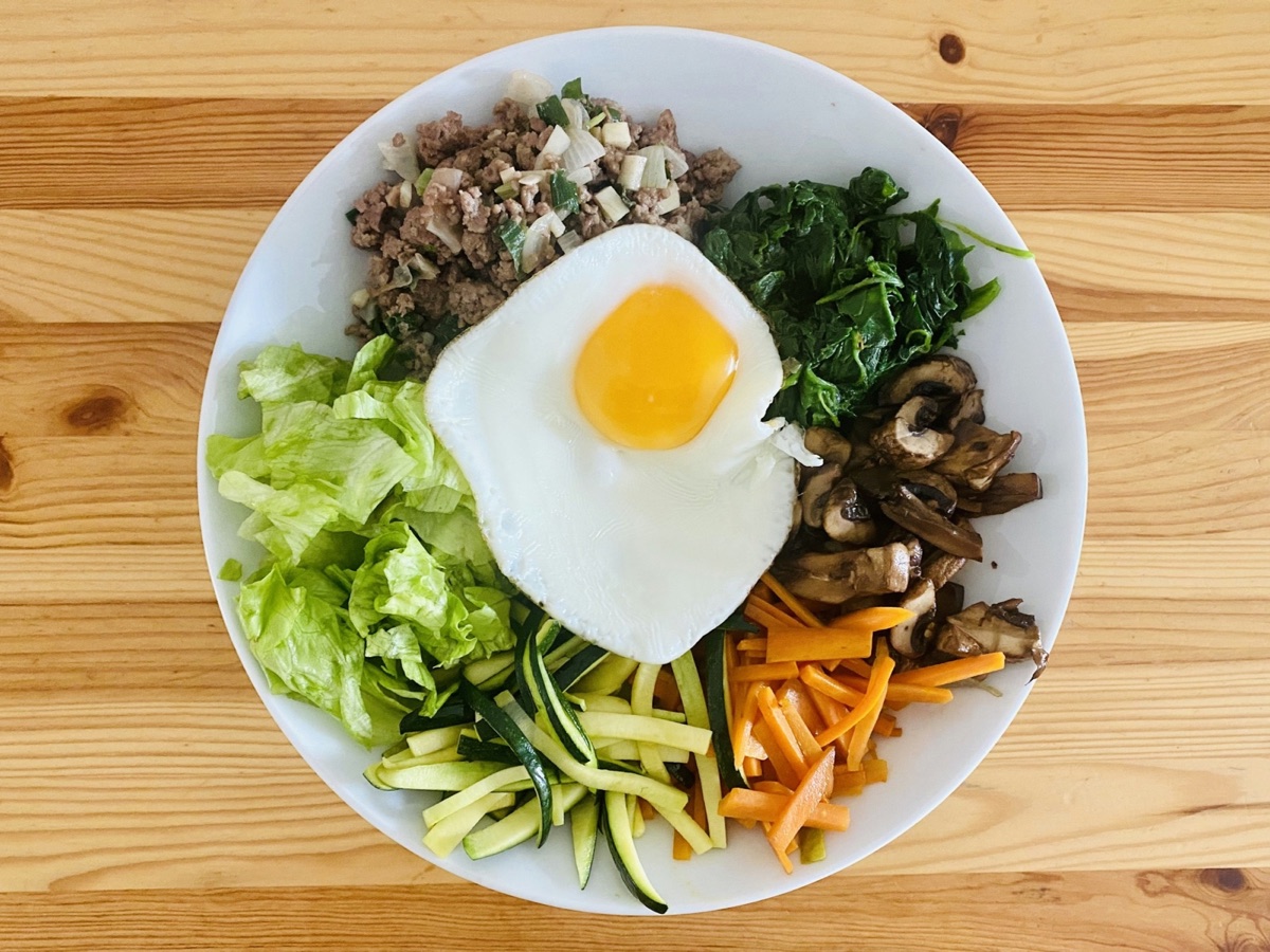 Symbolfoto: Bibimbap auf einem weißen Teller mit Spinat, Pilzen, Möhre, Zucchini, Salat, Hackfleisch und Spiegelei