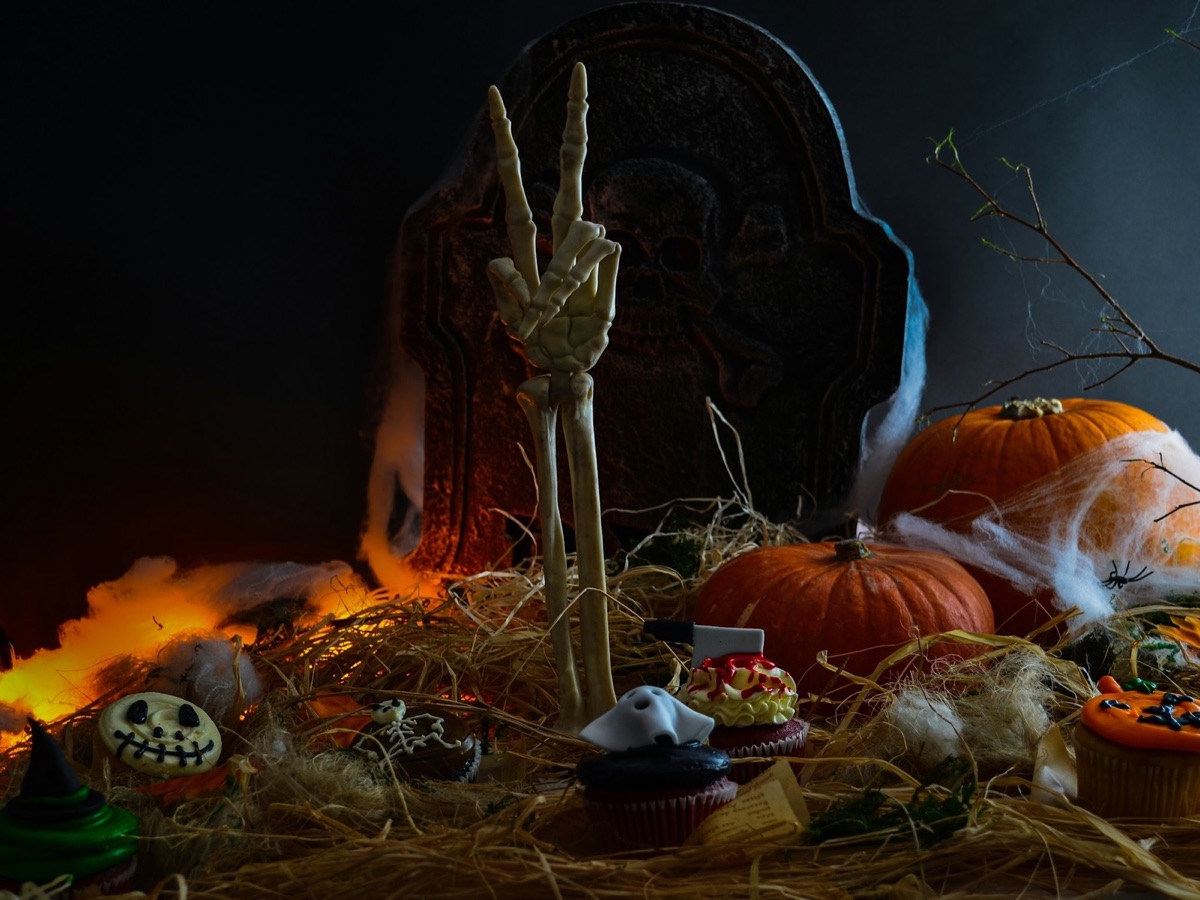 Symbolfoto: Eine Skeletthand ragt vor einem Grabstein aus dem Stroh, ringsum Kürbisse und Halloween-Gebäck